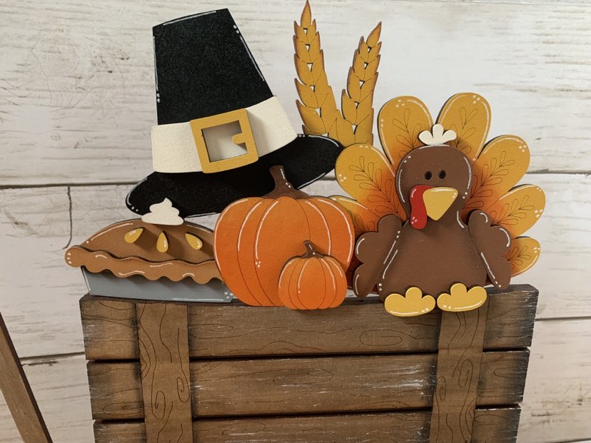 A Thanksgiving-themed basket insert featuring a turkey, pumpkin, and a pilgrim hat, all handpainted and placed in a wooden basket.