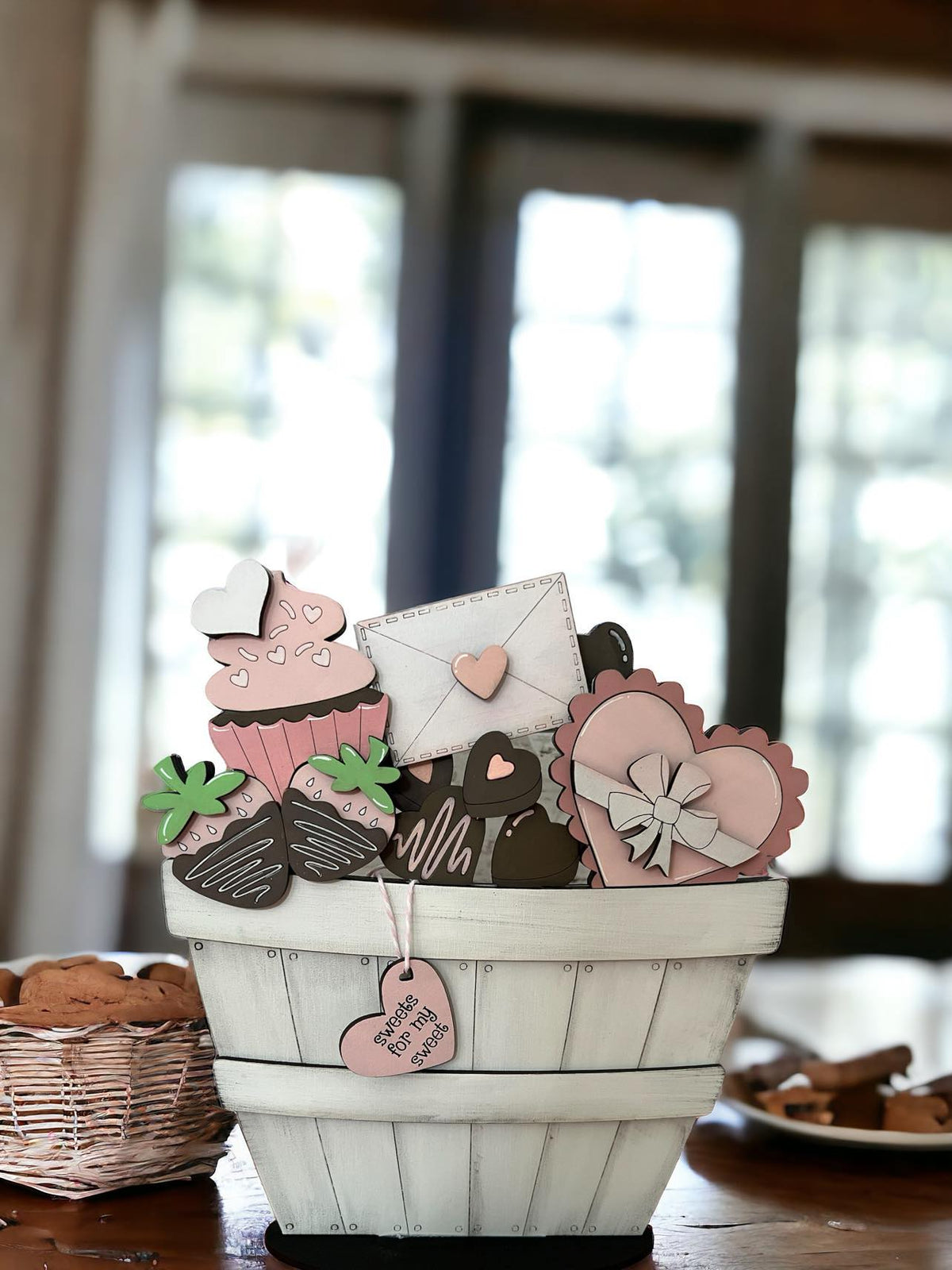 A decorative basket with handpainted and laser-cut items including hearts, chocolates, and a cupcake, all in shades of pink, white, and brown, placed on a wooden surface with a blurred background.