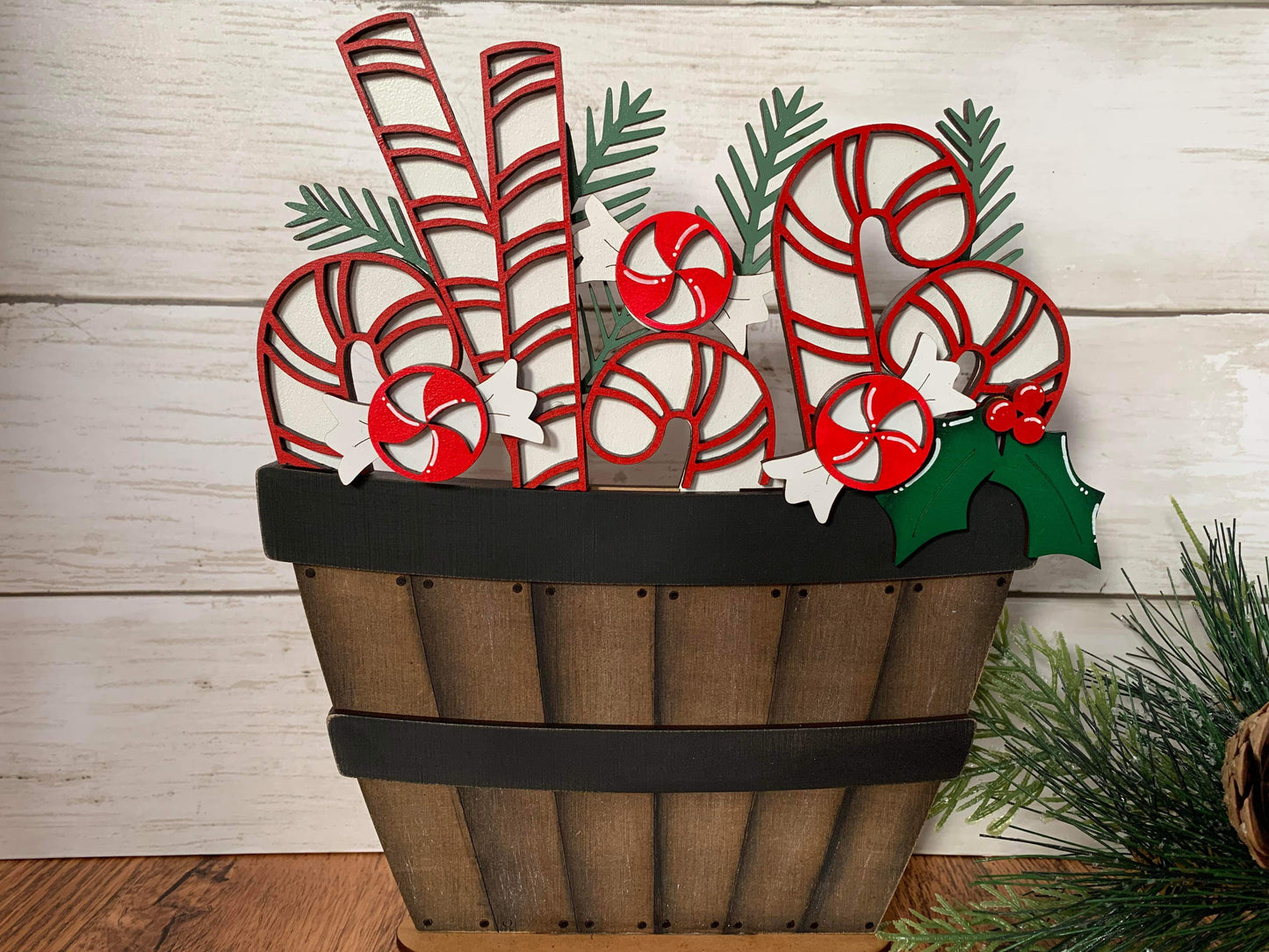 A decorative basket with a Christmas candy cane theme, featuring peppermint candy cane shapes in a brown insert, placed beside a wooden background with greenery.
