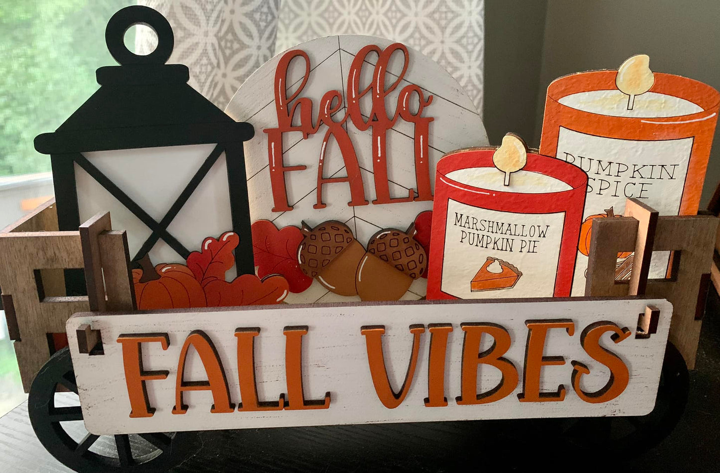 A decorative fall-themed wagon insert with the text 'FALL VIBES' on a white board, accompanied by a black lantern, a red candle with 'PUMPKIN SPICE' text, and wooden pieces shaped like leaves and a pumpkin.