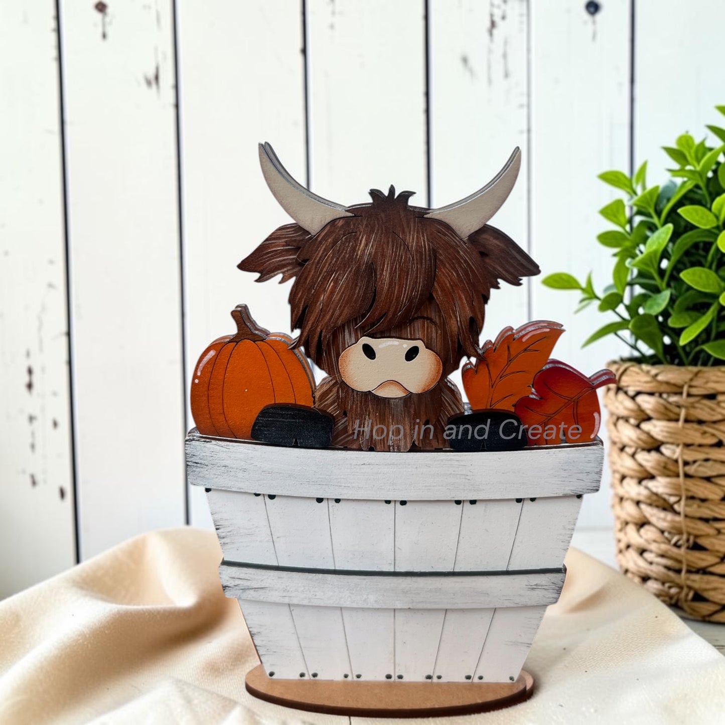 A decorative Highland cow figurine with long brown hair and curved horns, sitting inside a white wooden crate with pumpkins and autumn leaves, displayed on a beige fabric surface with a white wood plank background and a potted plant.
