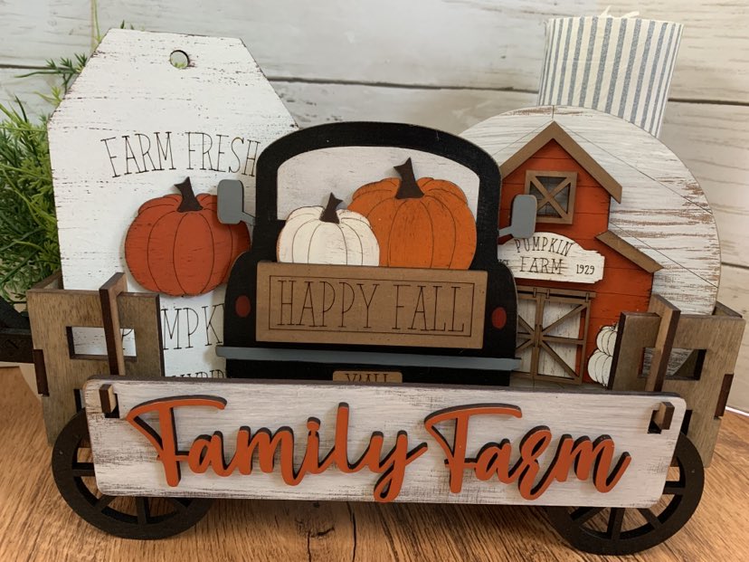 A seasonal decoration featuring a wooden farmhouse with a hayride, filled with pumpkins, and text that reads 'Fall Family Farm'.