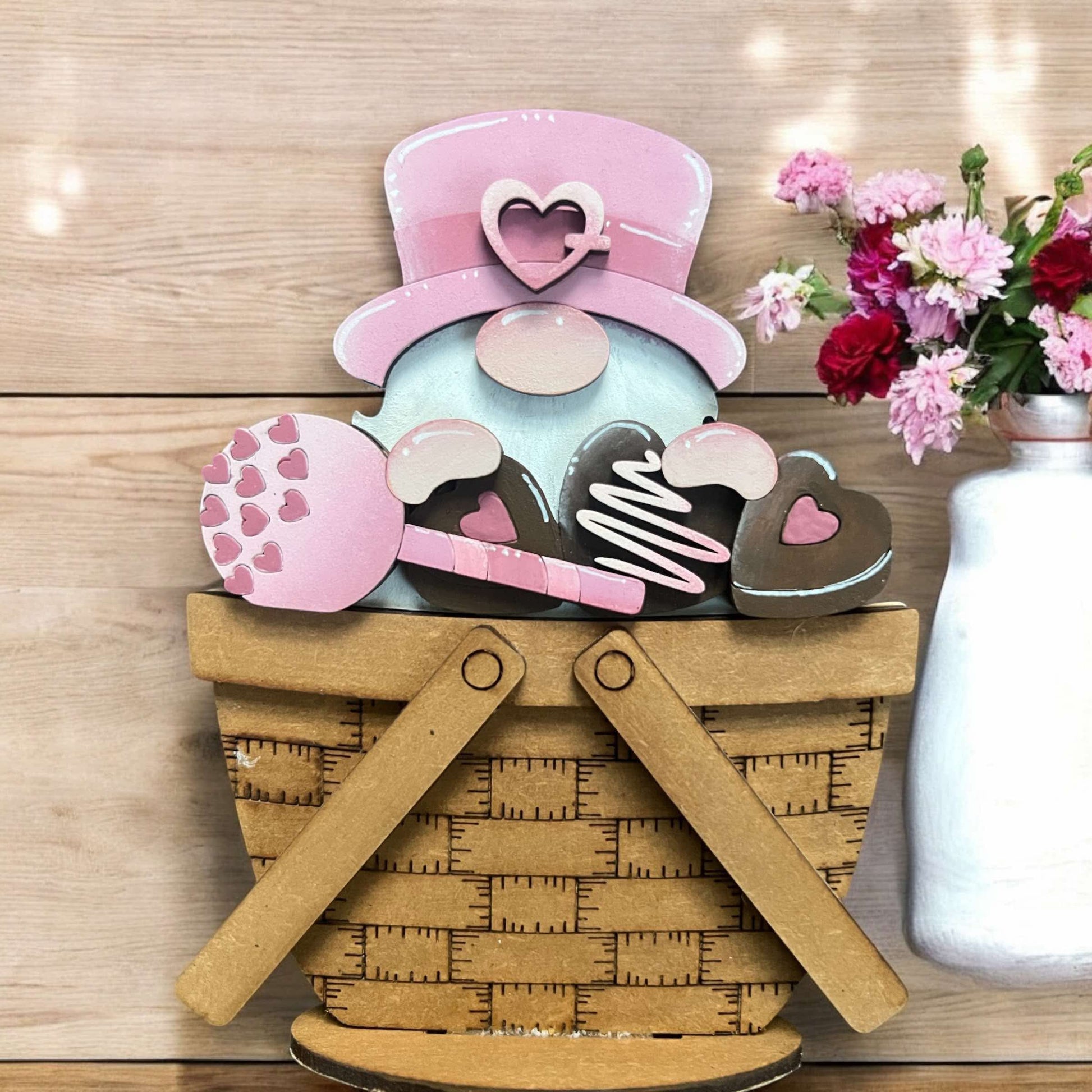 A mini Valentine gnome standing in a basket with a pink hat, heart-shaped cookies, and a flower, against a backdrop of a wooden surface and pink flowers.