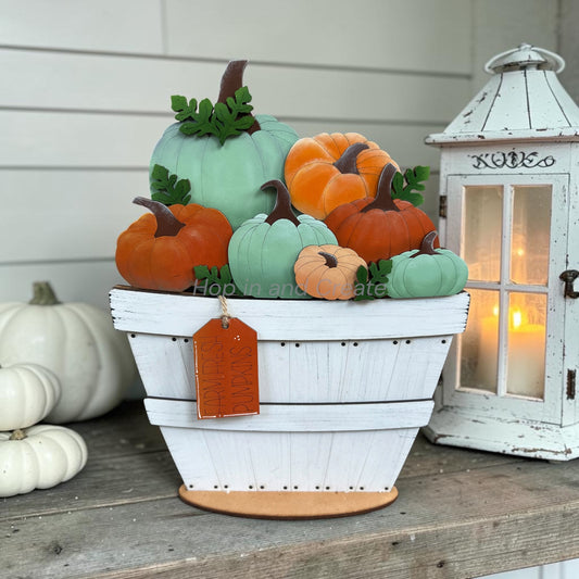 A decorative autumn pumpkin arrangement in a white wooden basket with a hanging orange tag, displayed on a rustic wooden surface beside a white lantern and small white pumpkins.