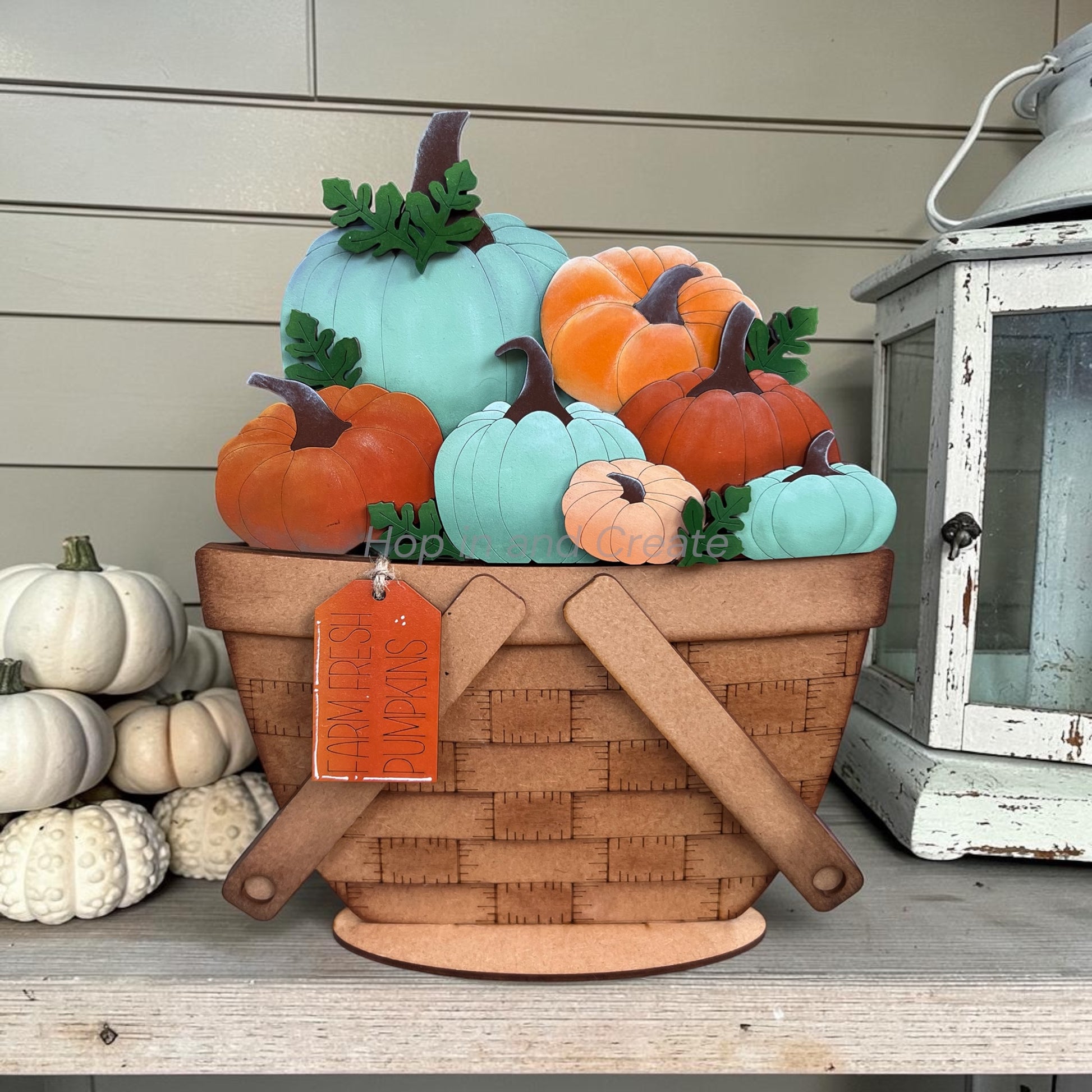 A decorative autumn pumpkin basket arrangement on a shelf, featuring a woven brown basket filled with pastel pumpkins in mint, orange, coral, and peach, with faux green leaves and stems. A hanging orange tag is attached to the basket, and a white lantern and stacked white pumpkins are visible in the background.