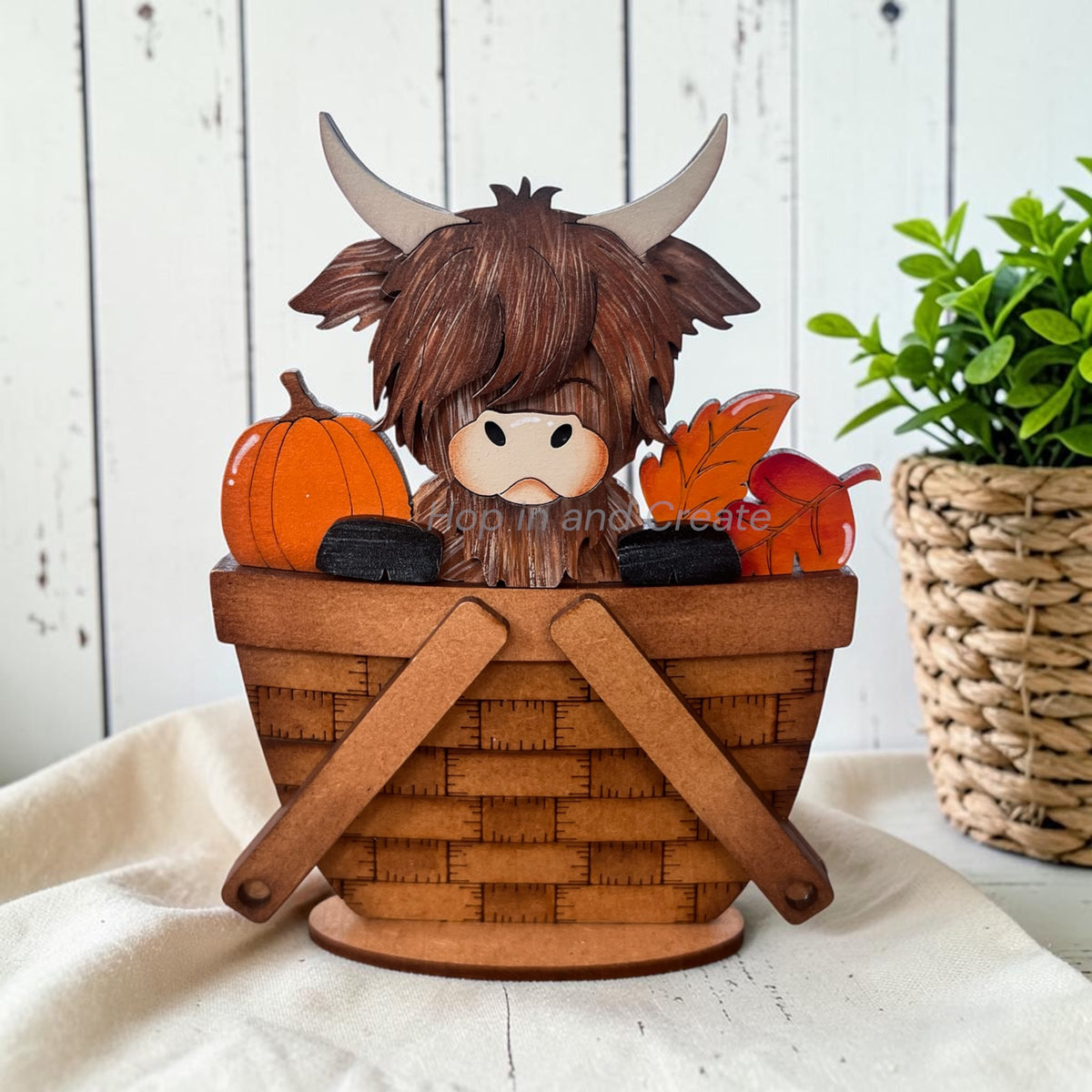 A decorative Highland cow figurine sitting in a woven basket, surrounded by autumn pumpkins, leaves, and black stones, displayed on a white fabric surface with a rustic white wood background and a potted plant.