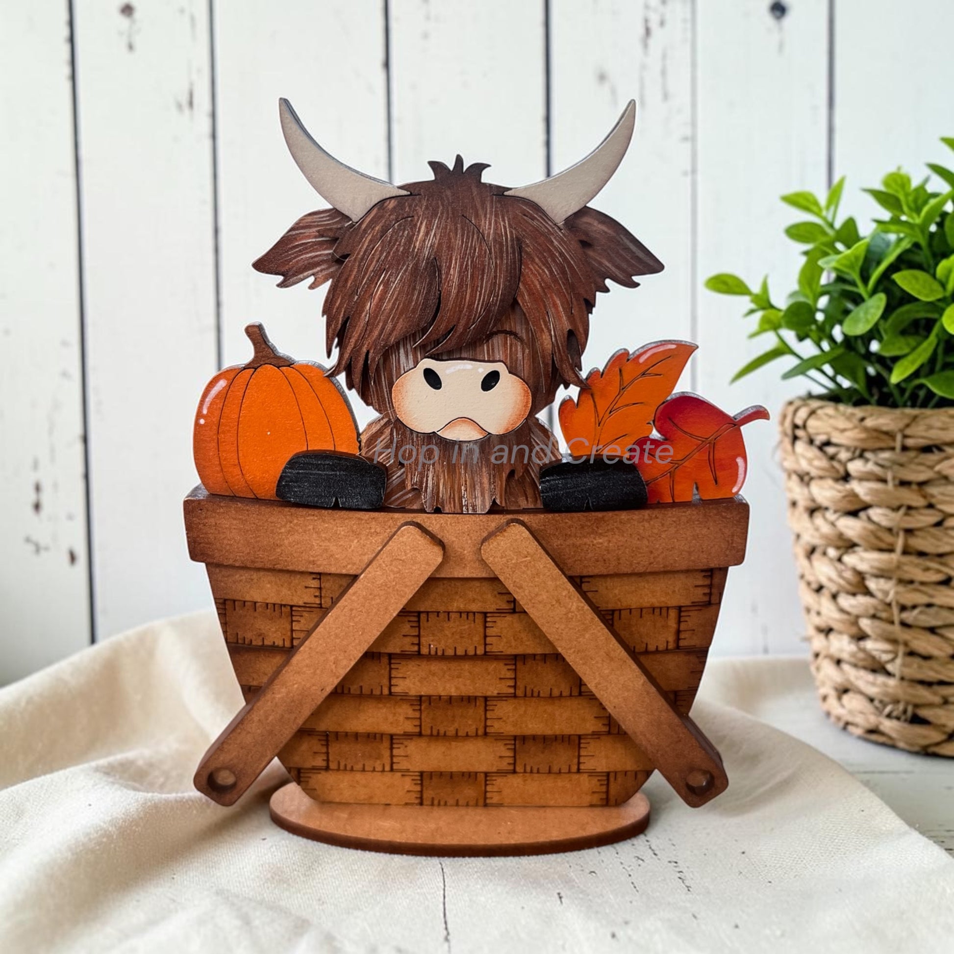 A decorative Highland cow figurine sitting in a woven basket, surrounded by autumn pumpkins, leaves, and black stones, displayed on a white fabric surface with a rustic white wood background and a potted plant.