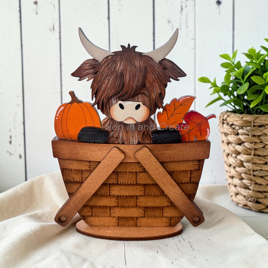 A decorative Highland cow figurine sitting in a woven basket, surrounded by autumn pumpkins, leaves, and black stones, displayed on a white fabric surface with a rustic white wood background and a potted plant.