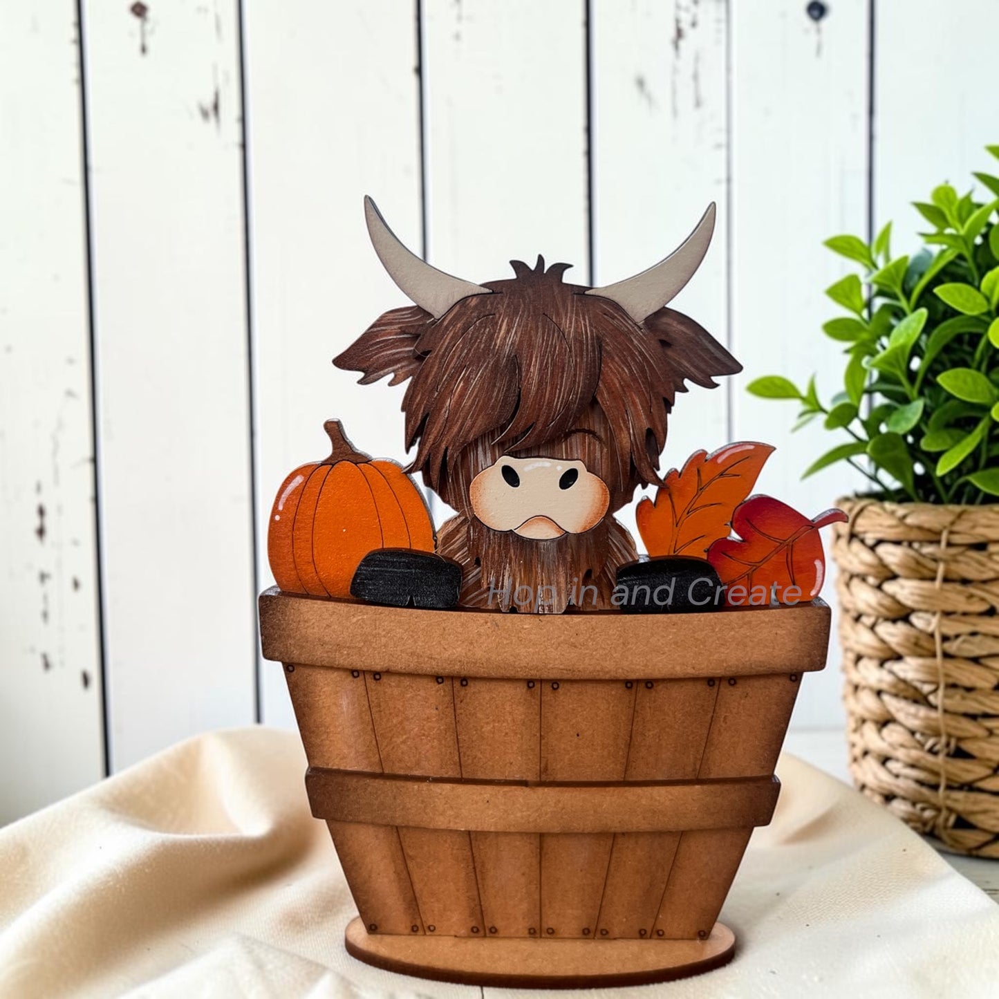 A decorative wooden Highland cow figurine sitting in a small brown basket, surrounded by orange pumpkins and autumn leaves, displayed on a cream fabric surface with a white wood plank wall and a potted plant in the background.