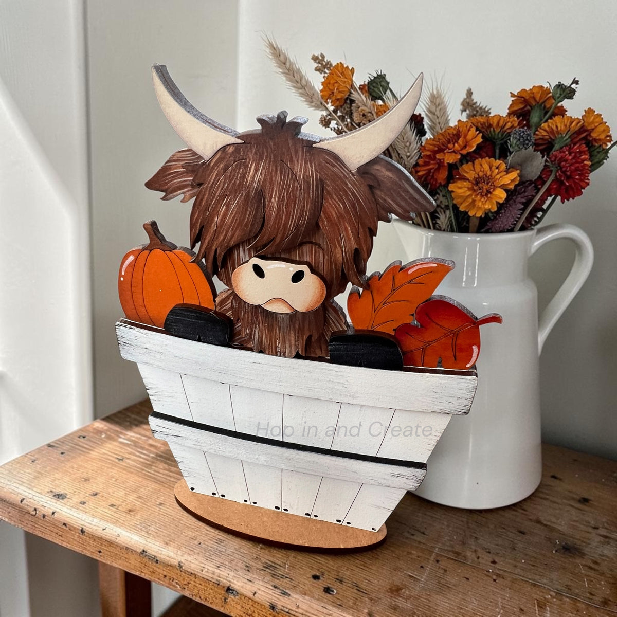 A decorative Highland cow figurine with brown shaggy hair and cream horns, sitting in a white wooden bucket filled with pumpkins, orange leaves, and black tires, displayed on a rustic wooden table next to a white pitcher of dried flowers.