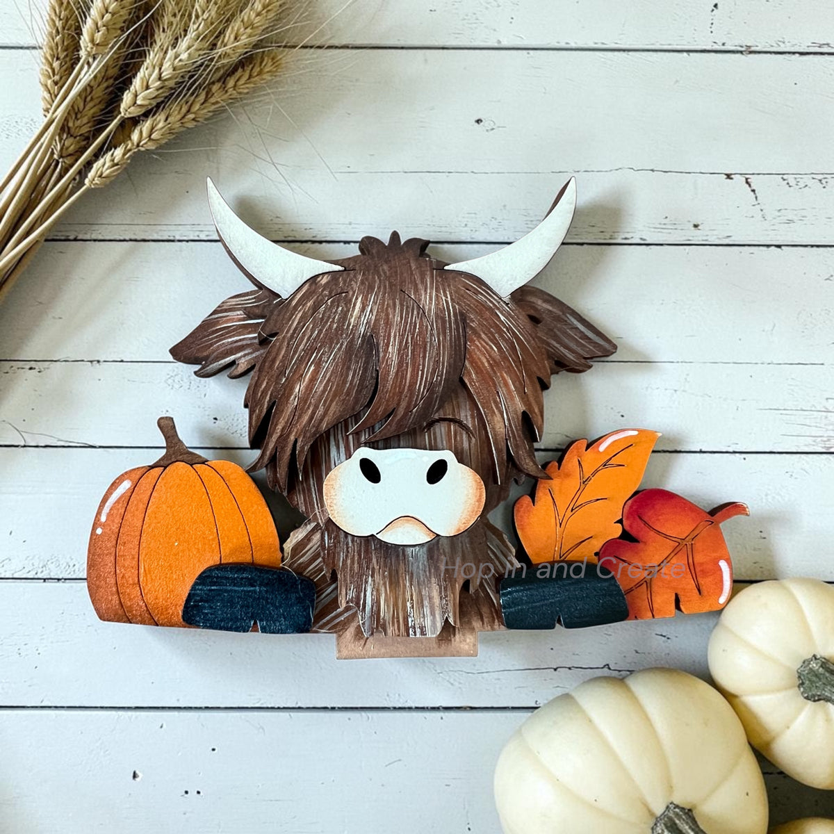 A rustic wooden Highland cow wall decor piece with white horns, shaggy brown hair, and a white snout, styled with autumn pumpkins, leaves, and wheat on a white wood plank background.