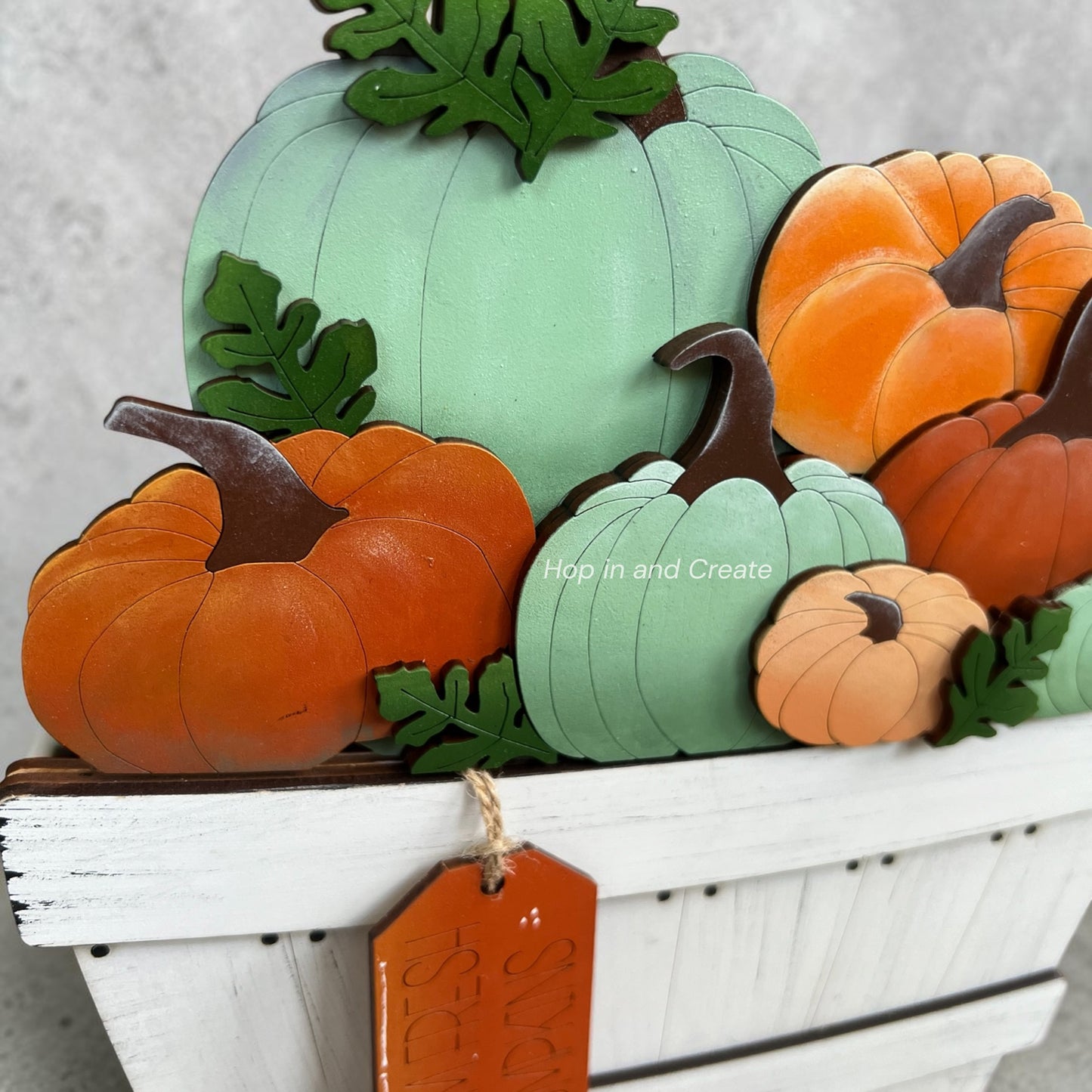 A decorative autumn pumpkin arrangement featuring multiple faux pumpkins in orange, mint green, peach, and brown, styled in a white wooden basket with green leaves and a hanging orange tag.