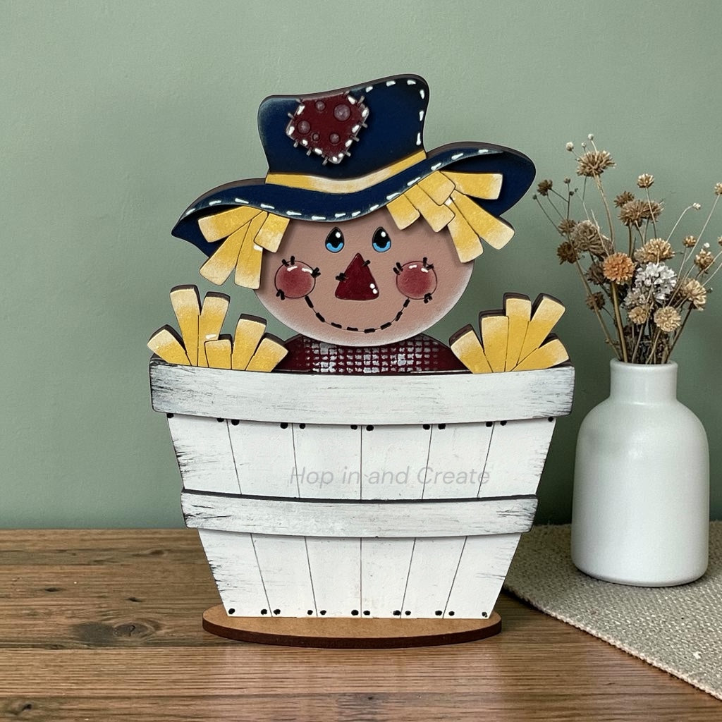 A rustic wooden decorative scarecrow sitting in a white painted barrel, displayed on a wooden tabletop next to a white vase with dried flowers against a sage green wall.