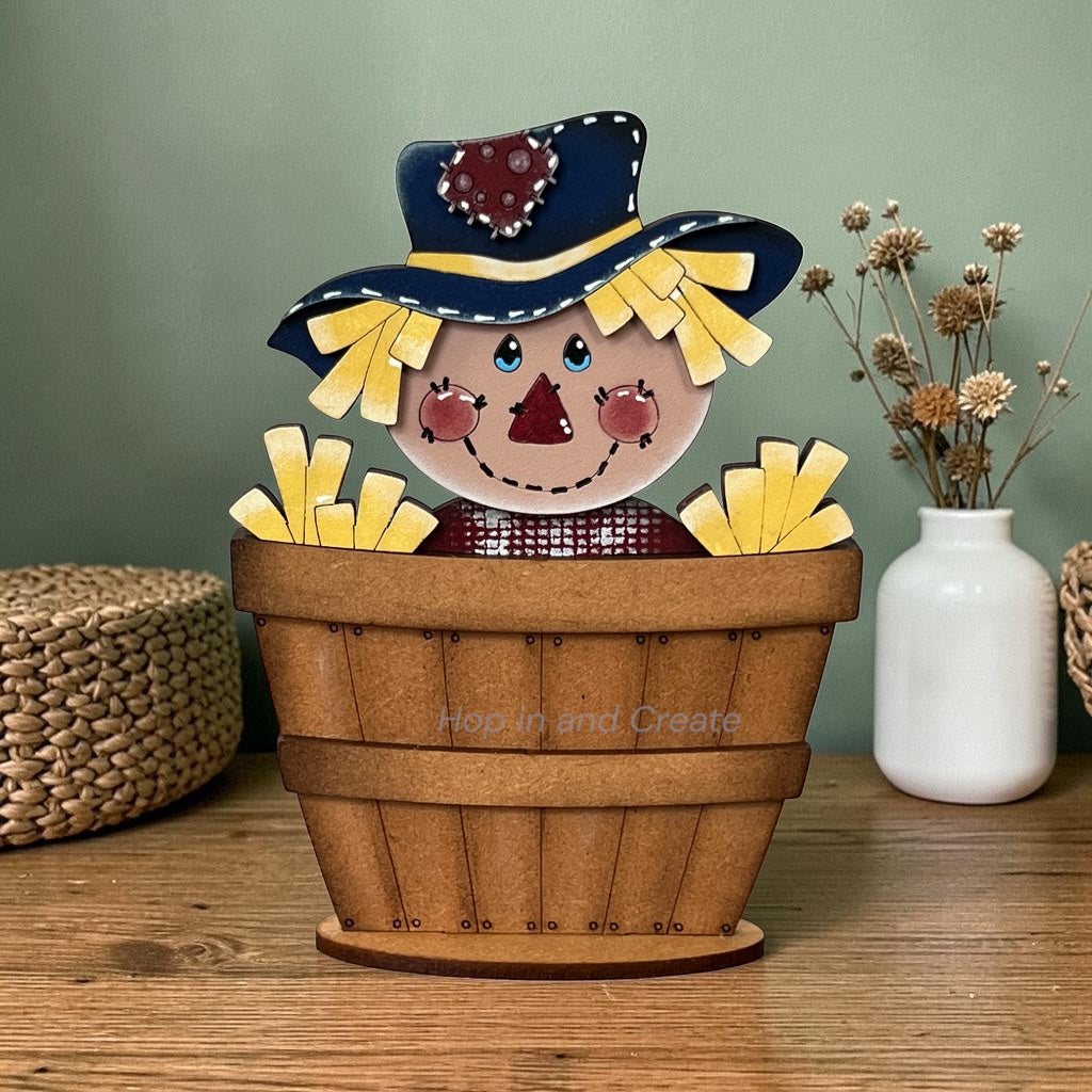 A decorative wooden scarecrow in a brown basket, displayed on a wooden tabletop against a sage green wall, with a white vase of dried flowers and woven baskets in the background.