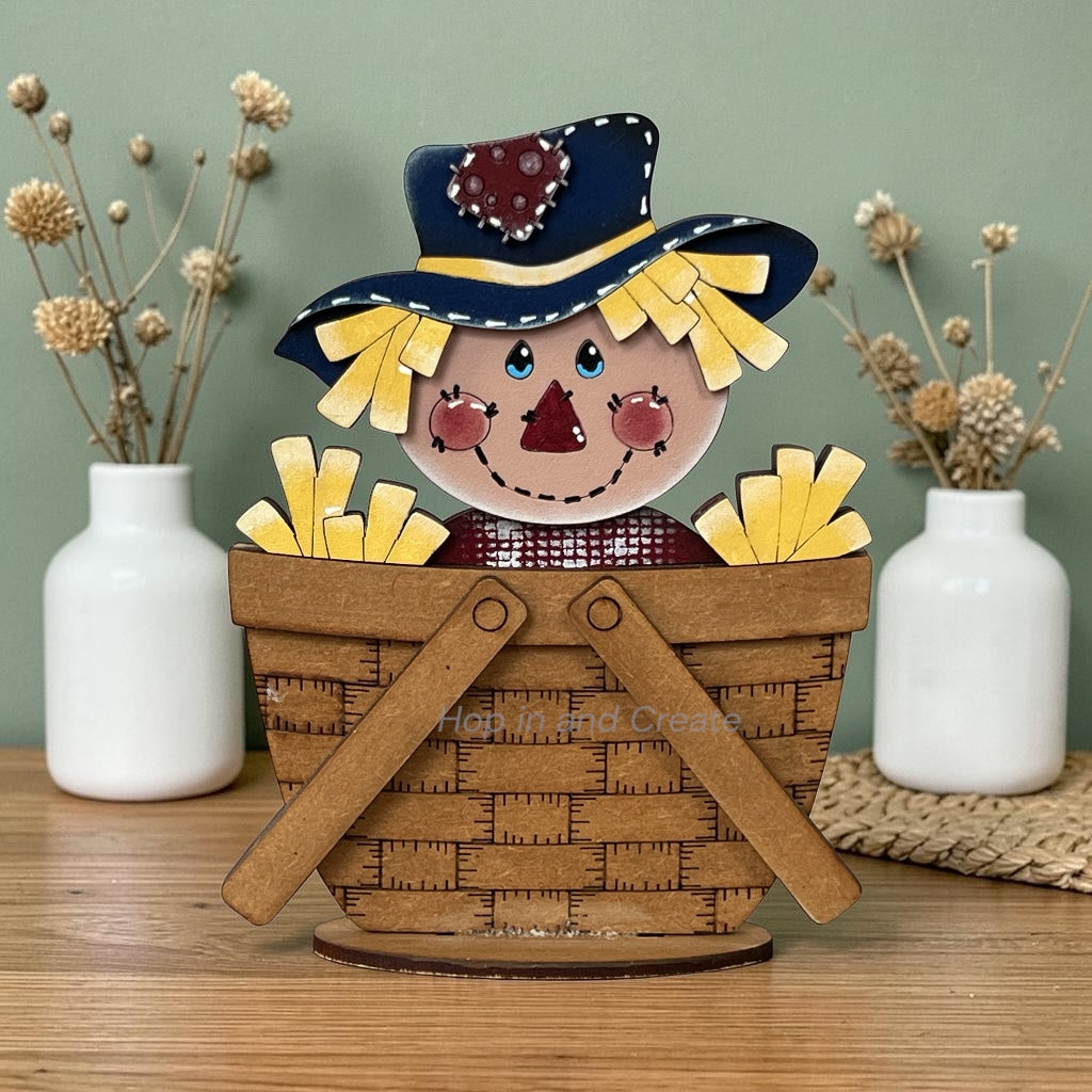 A decorative wooden scarecrow in a woven basket, styled on a wooden tabletop with two white ceramic vases containing dried flowers against a sage green background.