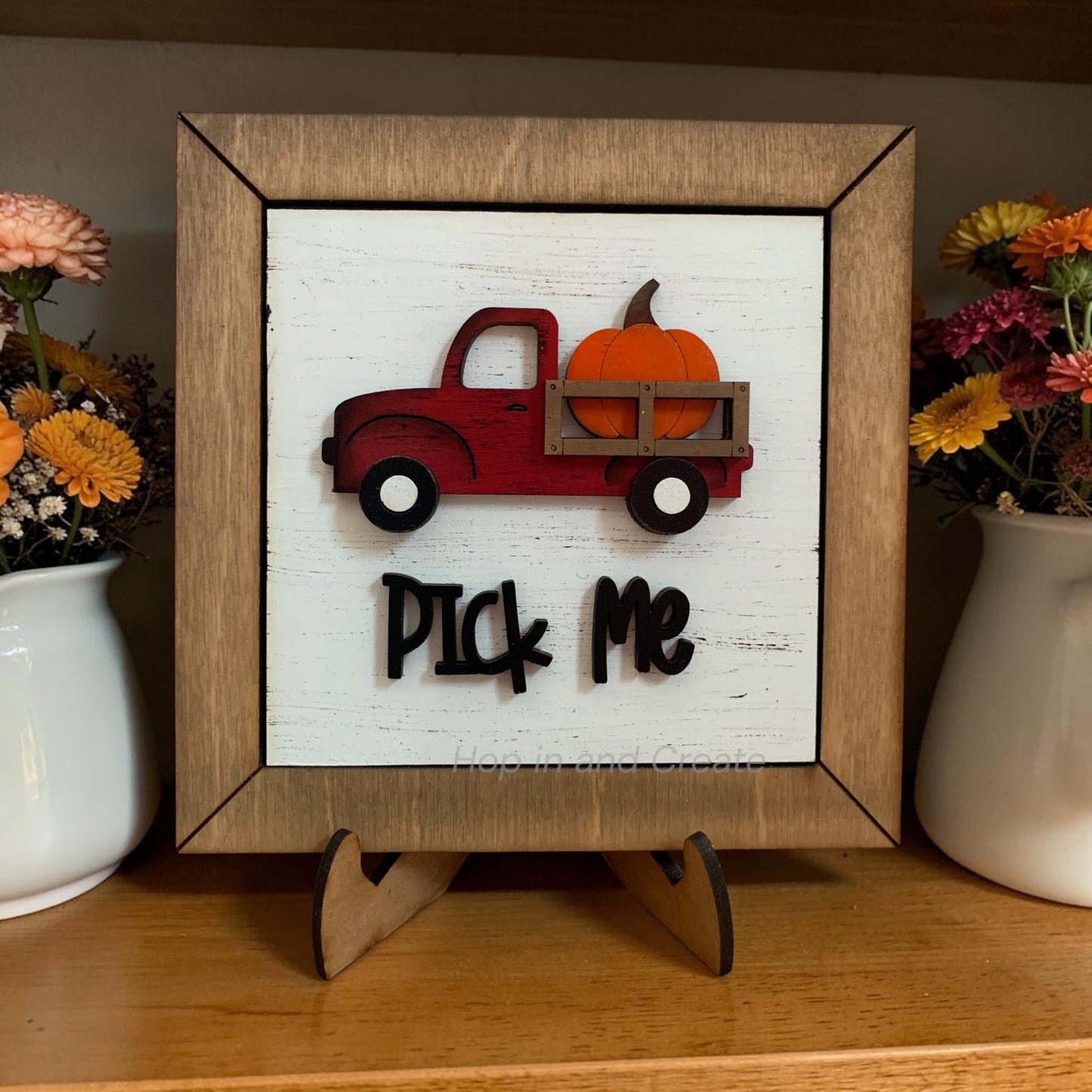 Decorative sign with a red truck and pumpkin on a white background, framed in wood, placed on a wooden surface with flowers.