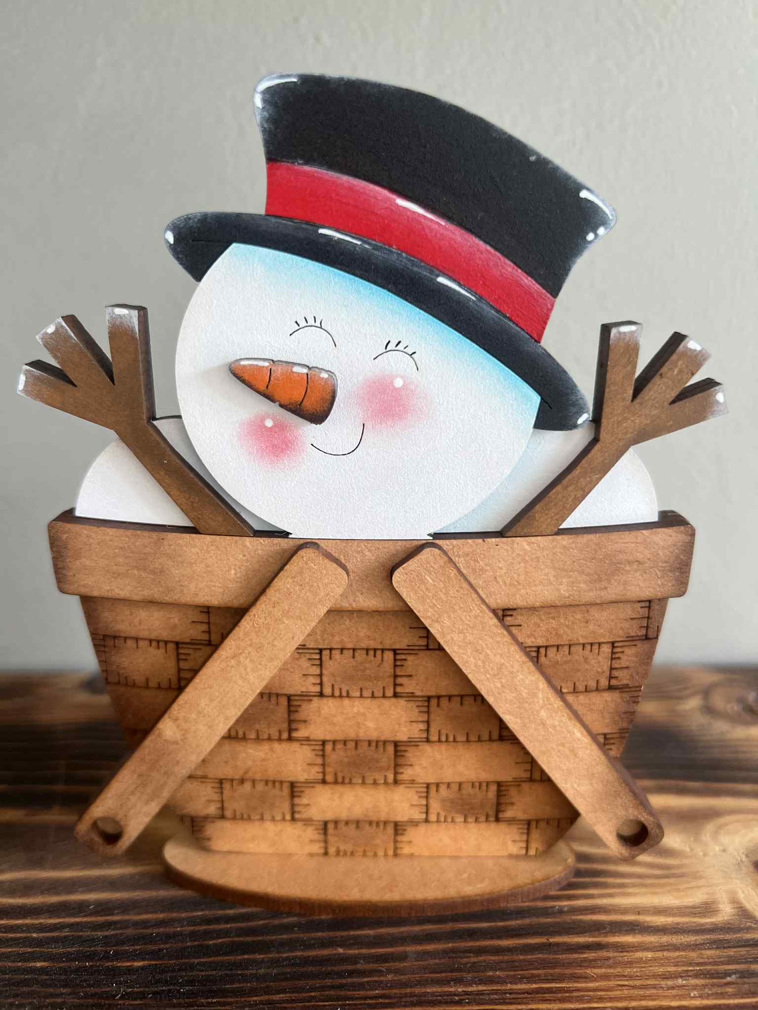 Decorative snowman figurine with a black top hat and carrot nose inside a wooden basket on a wooden surface.