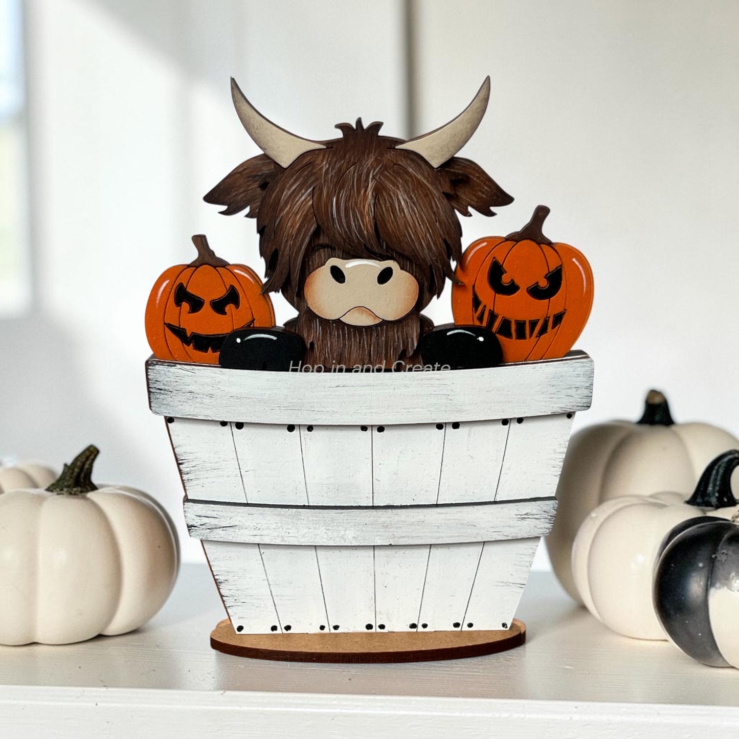 A decorative Highland cow figurine sitting inside a white wooden bucket planter, surrounded by orange jack-o'-lantern pumpkins, displayed on a white shelf with additional pumpkins in the background.