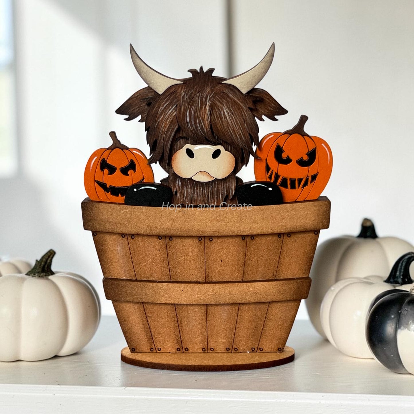A decorative Highland cow figurine sitting inside a wooden bucket with two orange jack-o'-lantern pumpkins behind it, displayed on a white surface with small white and black pumpkins in the background.
