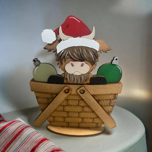 A decorative mini basket insert featuring a highland cow with a Santa hat and green Christmas ornaments.