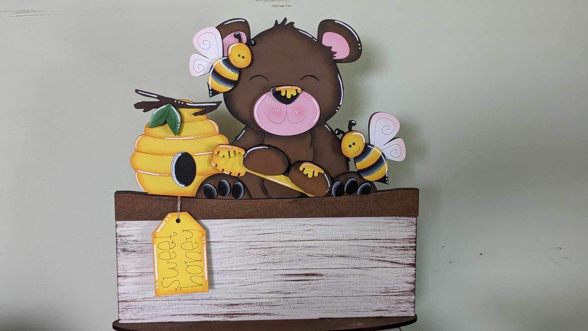 A decorative basket insert featuring a brown bear and bees around a honeycomb, with a large bee pollen tag, set against a beige background.