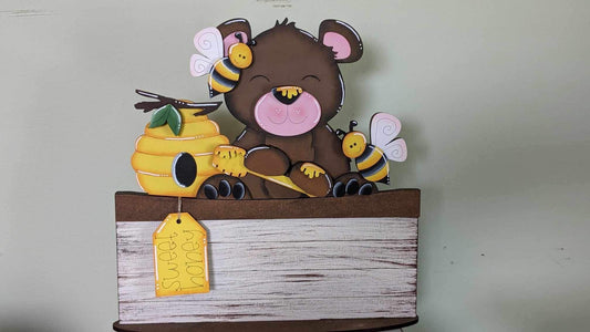 A decorative basket insert featuring a brown bear and bees around a honeycomb, with a large bee pollen tag, set against a beige background.