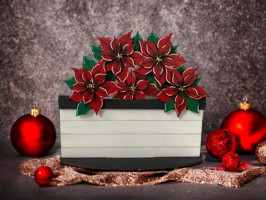 A decorative basket with a Christmas poinsetta design, featuring red and green poinsettias, placed on a glittery surface with Christmas ornaments around it.